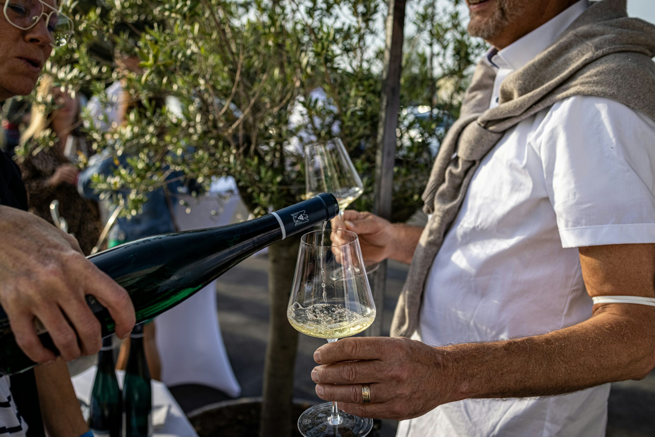 Mann in weißem Sommerhemd hält zwei Weingläser. Ein anderer schenkt Weißwein ein.